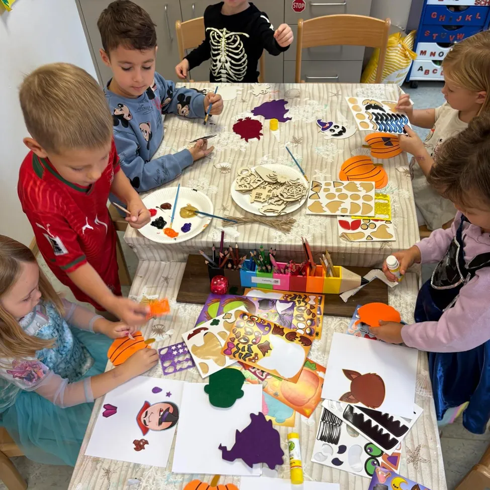 Eine Gruppe von Kindern ist um einen Tisch herum versammelt und beschäftigt sich mit einer Kunstaktivität. Sie verwenden Farbe, Aufkleber und andere Materialien, um Halloween-Themen-Basteleien zu erstellen.