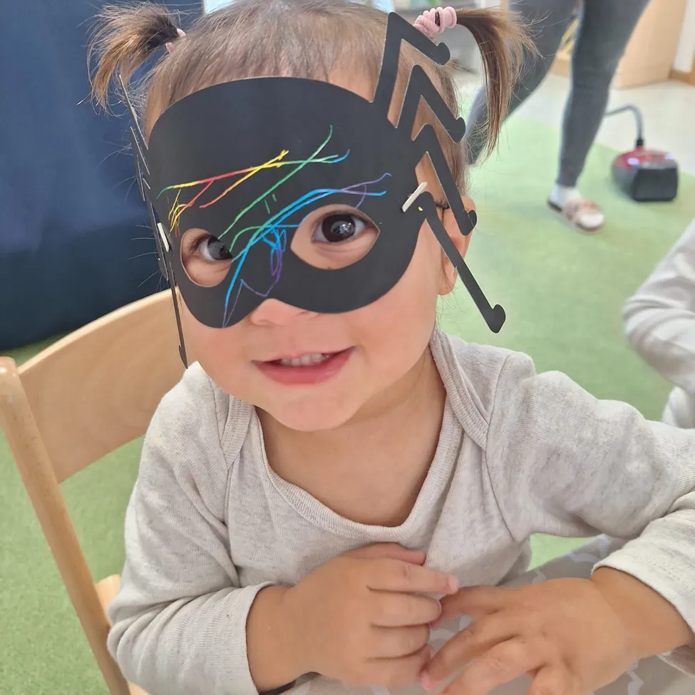 Ein kleines Mädchen trägt eine schwarze Spinnmaske mit bunten Zeichnungen darauf, sitzt auf einem Stuhl in einem Raum.