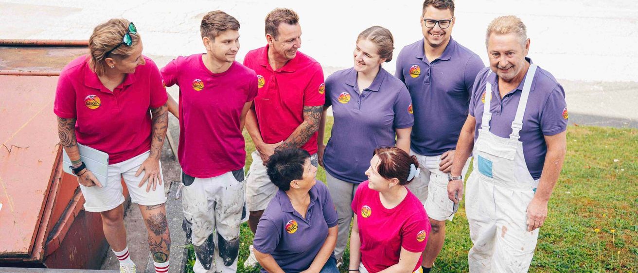 Eine Gruppe von Menschen in passenden Shirts steht und kniet auf Gras, lächelt und posiert für ein Foto mit einem Betonweg hinter ihnen.