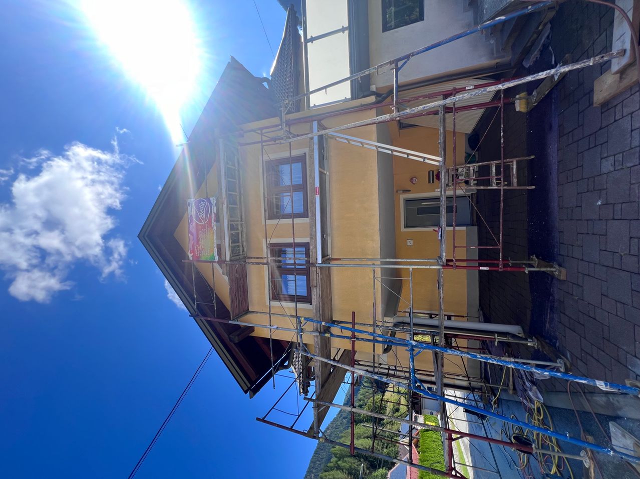 An aerial view of a yellow house under construction, with scaffolding surrounding it, a ladder, and a bright sun shining from the top left.