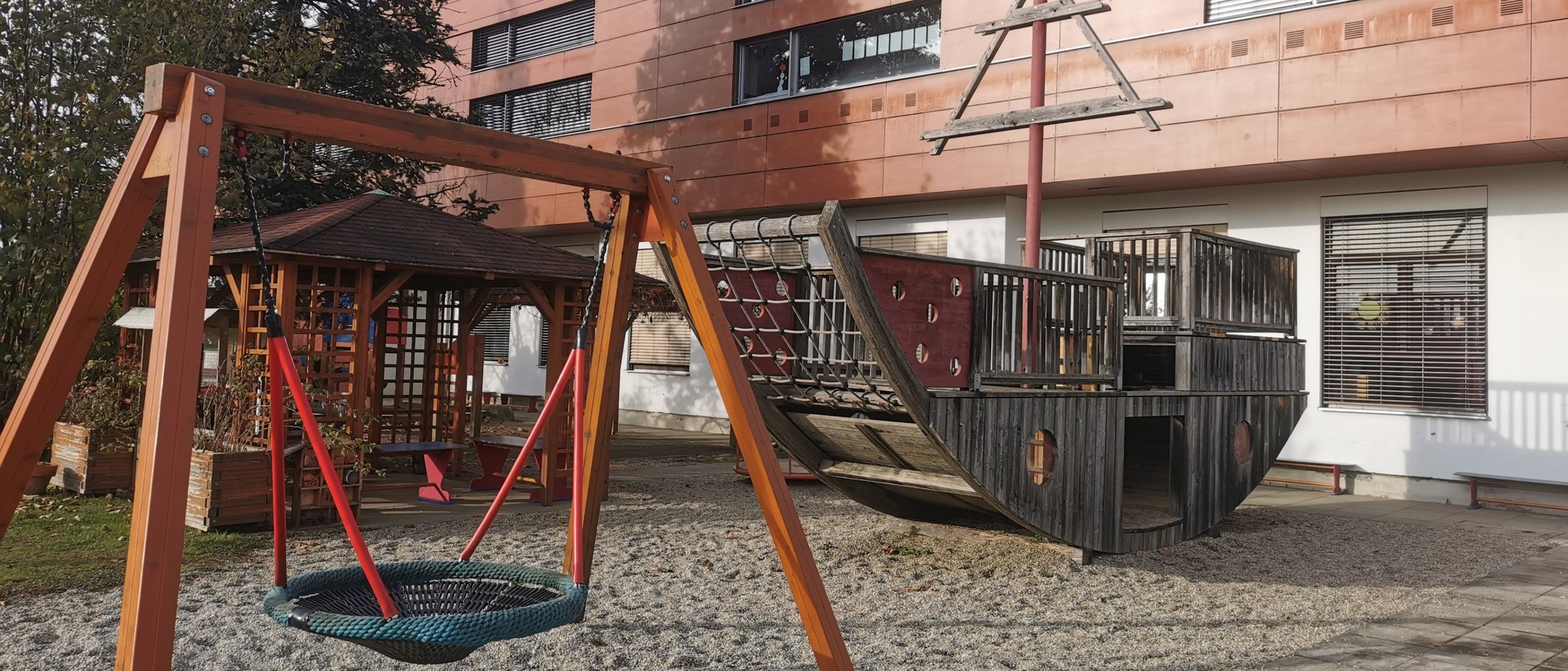 Ein Spielplatz mit einem hölzernen Piratenschiff und einem Schaukelset, umgeben von Kies und einem Gebäude mit Fenstern im Hintergrund.