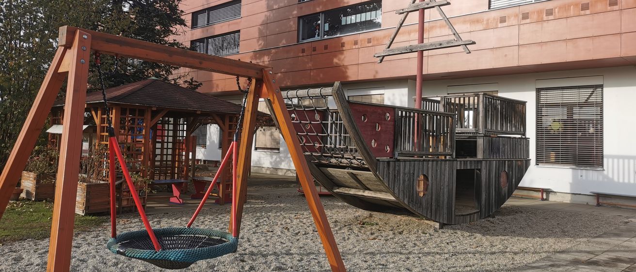 Ein Spielplatz mit einem hölzernen Piratenschiff und einem Schaukelset, umgeben von Kies und einem Gebäude mit Fenstern im Hintergrund.