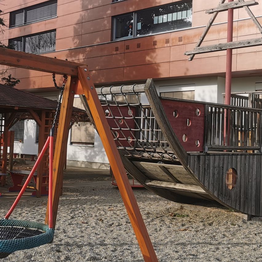 Ein Spielplatz mit einem hölzernen Piratenschiff und einem Schaukelset, umgeben von Kies und einem Gebäude mit Fenstern im Hintergrund.