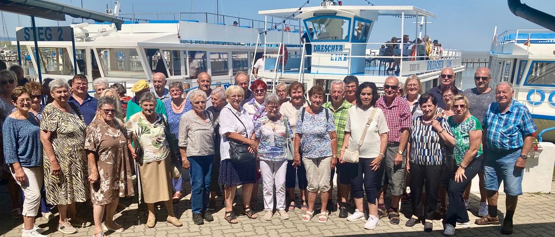Eine Gruppe älterer Menschen posiert für ein Foto am Hafen mit einem Fährschiff im Hintergrund.