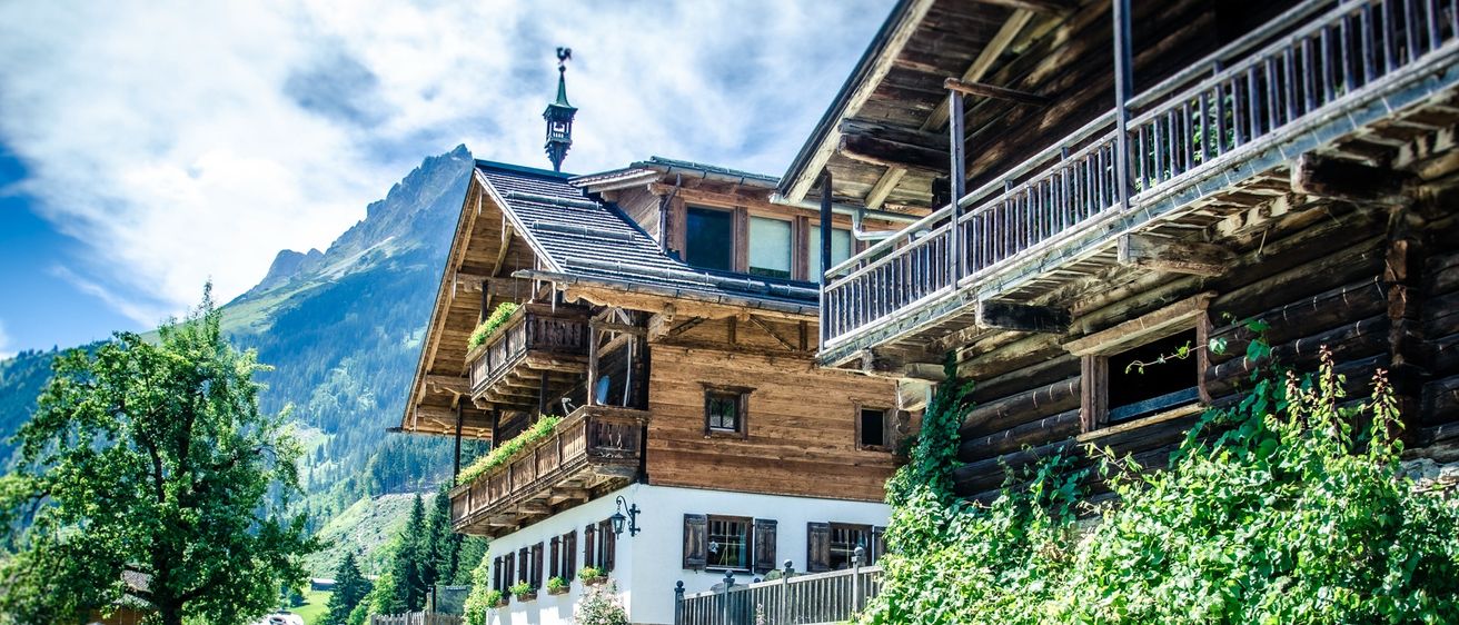 Ein Holzhaus mit Balkonen und einem Kirchturm, umgeben von Bergen, Grün und einem kleinen Holzzaun.