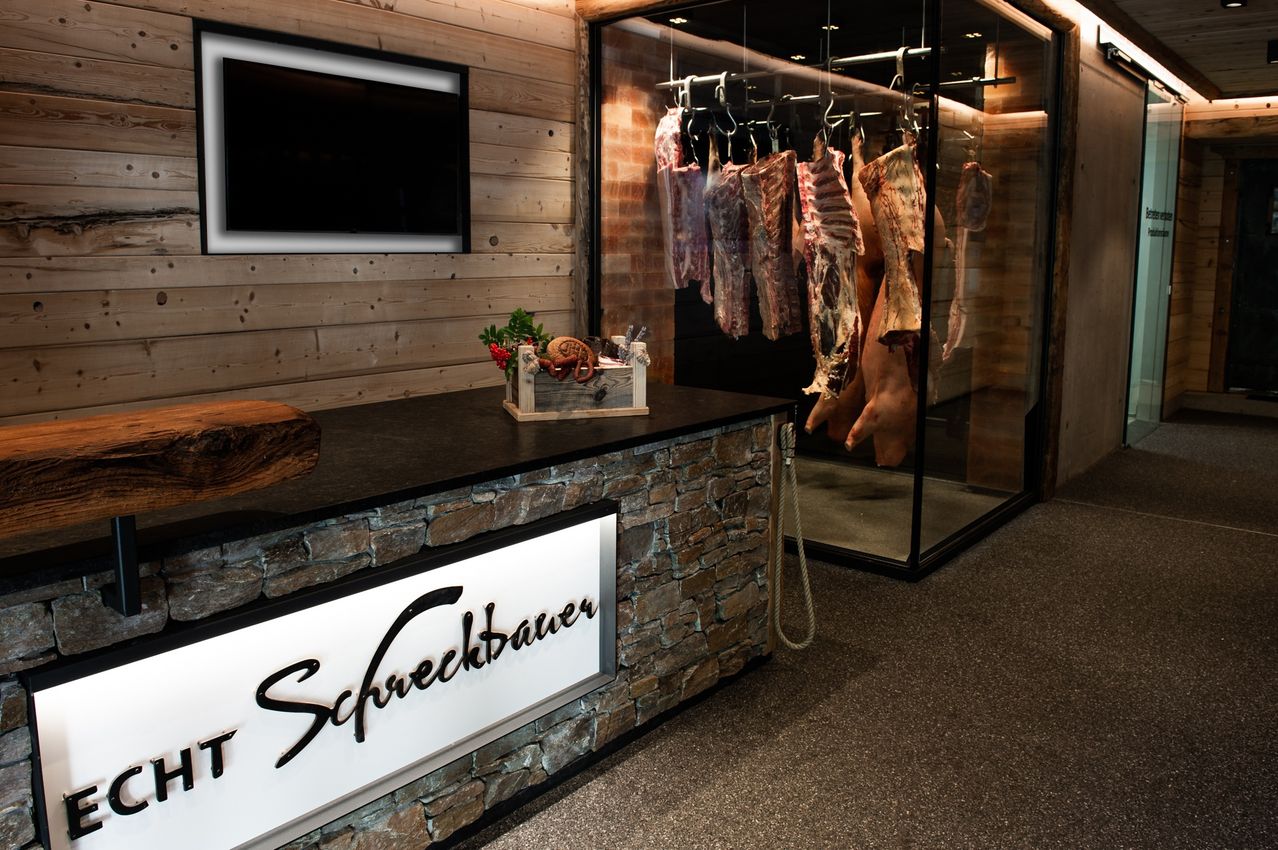 Ein Steintresen mit dem Wort 'Schreckbauer' darauf. Im Hintergrund befindet sich eine Glasvitrine mit hängendem Fleisch. Ein Fernseher ist an der Wand montiert.
