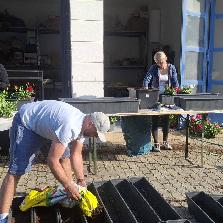 Ein Mann in einem weißen T-Shirt und blauen Shorts füllt mehrere schwarze Pflanzkübel mit Erde. Eine Frau in einer blauen Kapuzenjacke und einem weißen Oberteil steht an einem Tisch mit weiteren Pflanzkübeln.