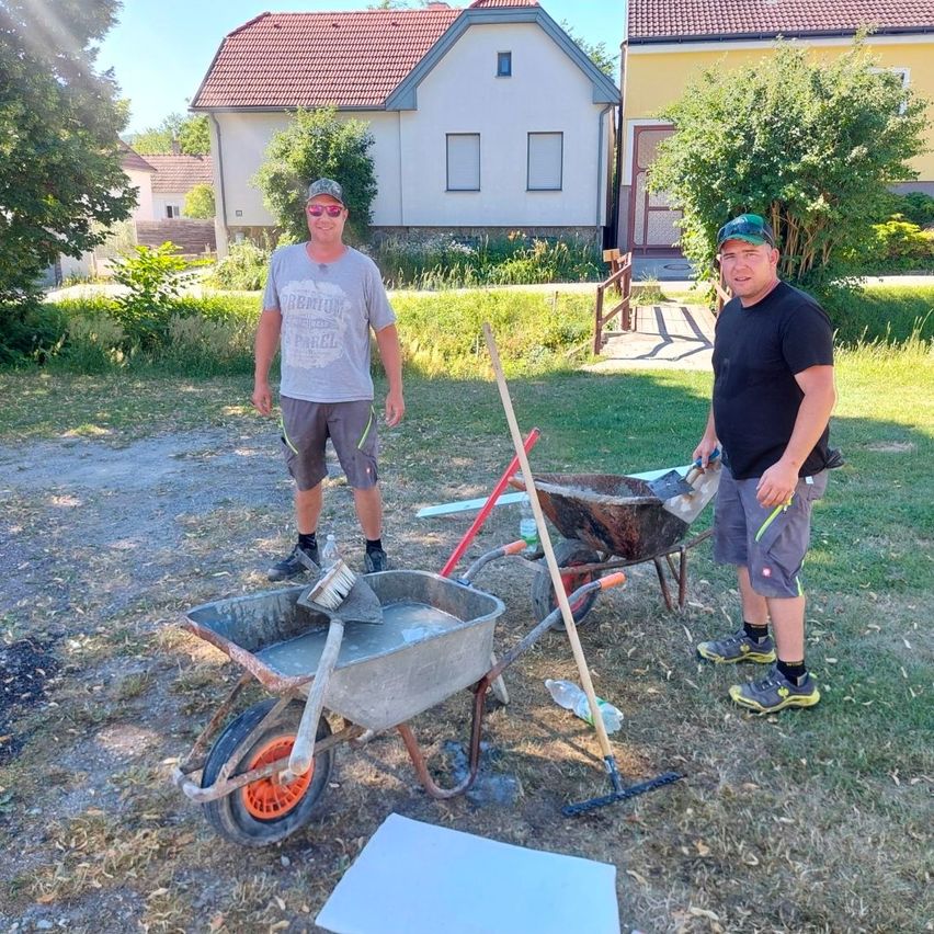 Zwei Männer stehen in einem Garten mit Werkzeug. Sie sind in der Nähe eines Schubkarrens und Schaufeln. Die Häuser sind hinter ihnen.