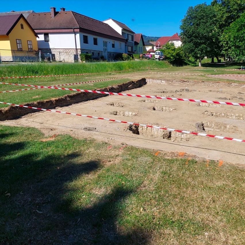 Ein archäologischer Ausgrabungsort ist von rotem und weißem Band umgeben. Hinter dem Gelände befinden sich Häuser und Bäume. Der Boden ist mit Erde bedeckt und einige Bereiche sind freigelegt.