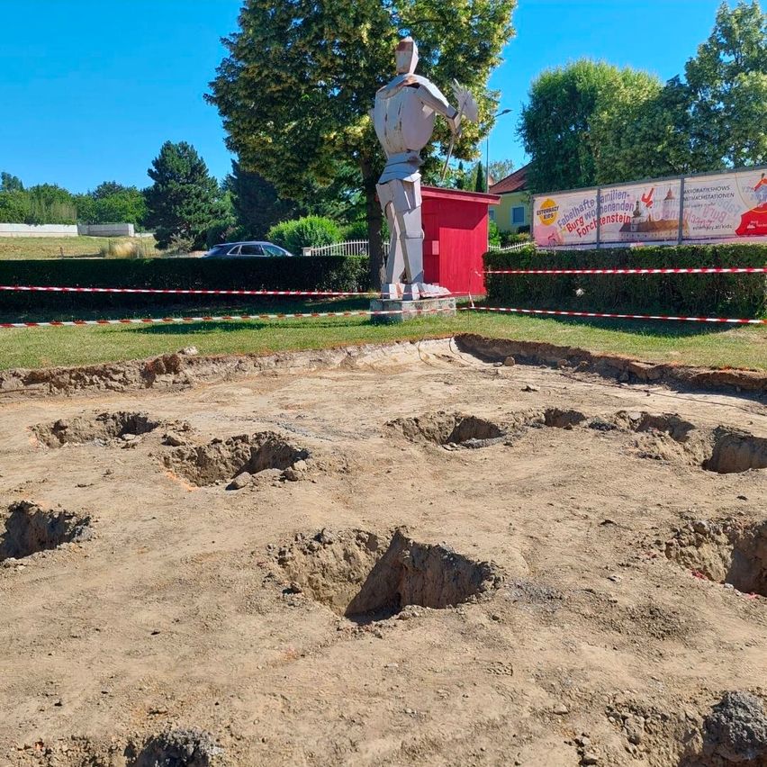 Eine Statue eines Ritters steht in der Nähe einer Baustelle mit ausgehobenem Boden, umgeben von roten und weißen Absperrbändern. Dahinter steht ein rotes Gebäude mit einer Fahne, und ein Auto ist in der Ferne geparkt.