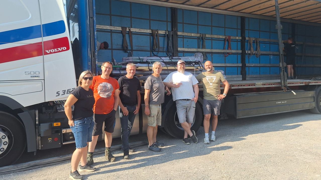Five individuals stand in front of a truck with various items loaded inside. The man on the left wears sunglasses, and the man in the middle wears a watch.
