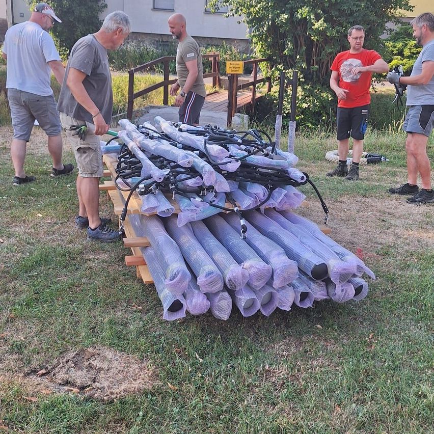 Five men are outside, two near a bridge and three near a pile of wrapped objects on a pallet. One man is gesturing.