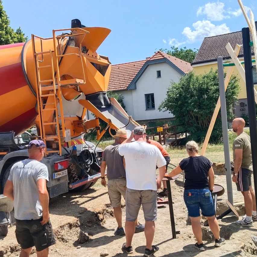 A concrete truck is pouring cement on a construction site. Several people stand around, including two men in hats and a woman in shorts. A house and trees are in the background.