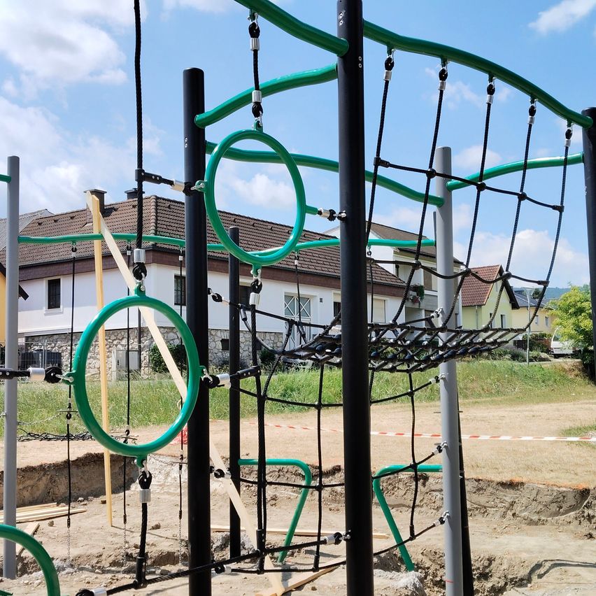 A playground with a large climbing frame, green and black colors, and rings hanging from it. The playground is under construction. There is a house in the background.