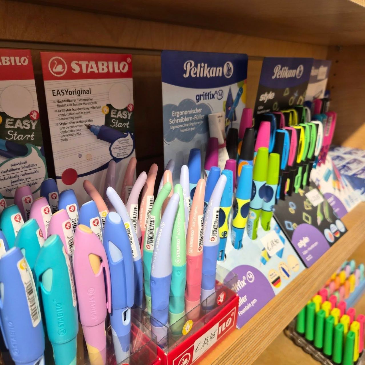 Ein Holzregal zeigt verschiedene farbige Stifte und Marker, einschließlich Stabilo und Pelikan. Die Stifte sind in verschiedenen Farben und Größen und einige sind mit verschiedenen Eigenschaften gekennzeichnet. Es gibt auch Schachteln mit Stiften und Markern auf dem Regal.