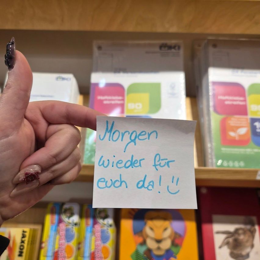 Eine Hand hält ein Stück Papier mit blauer Schrift, die 'Morgen wieder fur euch da!' besagt, vor einem Bücherregal. Der Hintergrund zeigt Bücher mit farbenfrohen Umschlägen.