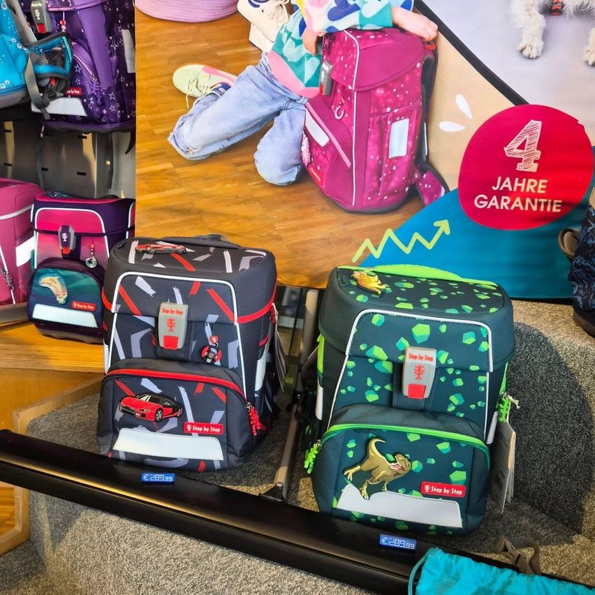 Verschiedene Kinderrucksäcke in unterschiedlichen Designs und Farben sind auf einer Holzoberfläche ausgestellt. Ein Poster im Hintergrund bewirbt eine 4-Jahres-Garantie.