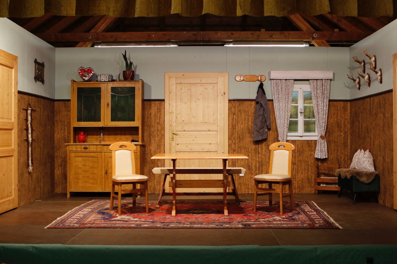 Eine einfache Bühneneinrichtung mit einem Holztisch und zwei Stühlen unter einer Holztür. Ein Schrank mit einer Pflanze und einem Herz darauf. Ein Fenster mit Vorhängen und einer hängenden Jacke.