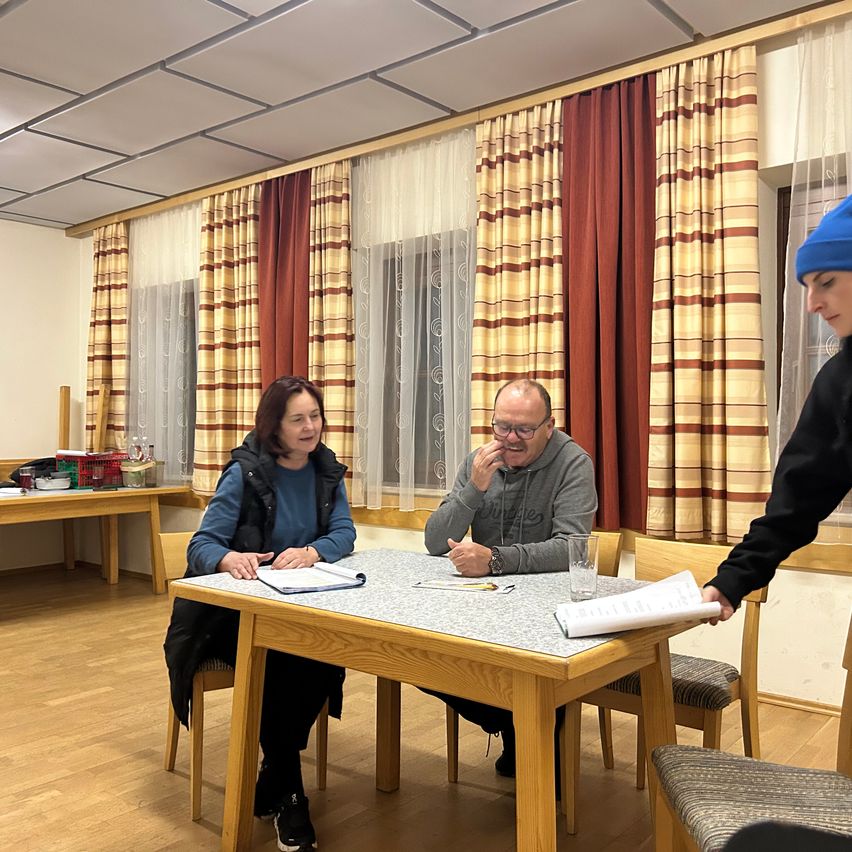 Ein Mann und eine Frau sitzen mit Büchern an einem Tisch und lächeln. Eine andere Person mit einer blauen Mütze steht neben ihnen. Der Raum hat Holzböden und einen Tisch mit Glasplatte. Es gibt Stühle, Vorhänge und einen Schreibtisch mit Gegenständen darauf.