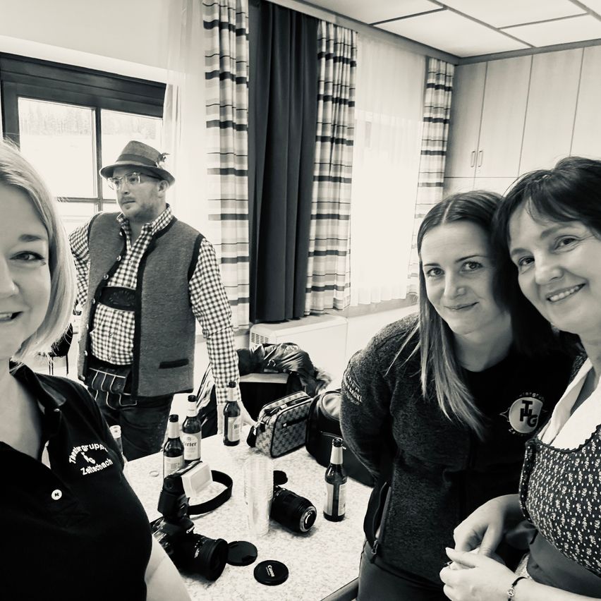 Drei Frauen und ein Mann lächeln in einem Raum. Die Frau links trägt eine Uniform mit Logo. Ein Tisch hat Flaschen und Kameras. Dahinter trägt ein Mann Hut und Weste.