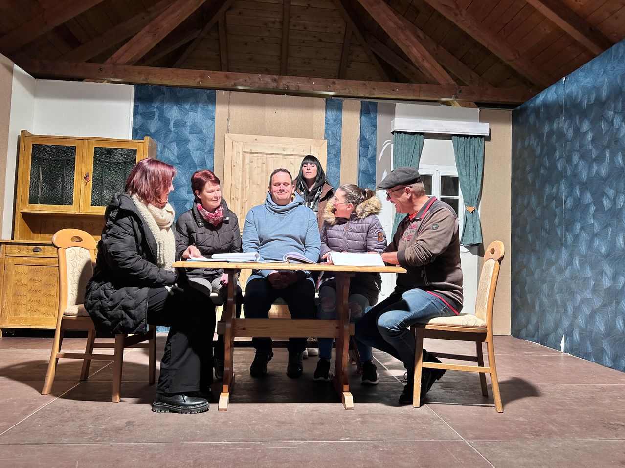 Sechs Personen sitzen um einen Holztisch herum, jeder hält ein Buch in der Hand. Sie scheinen eine Theaterprobe zu üben, wie ihre Kleidung und die Umgebung vermuten lassen.