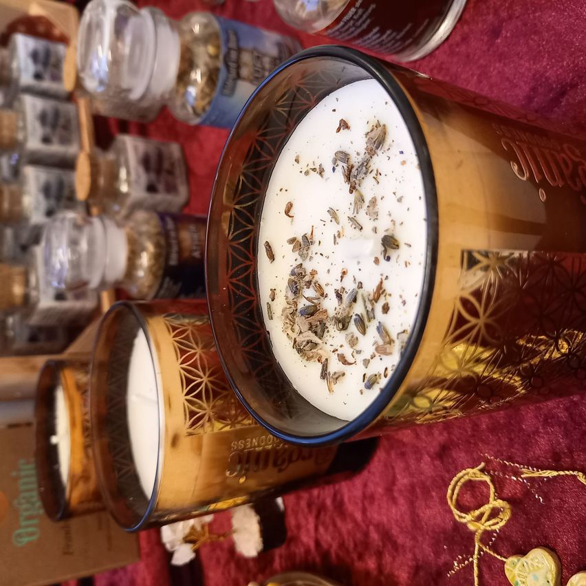 Ein Nahaufnahme eines Kerzenbechers mit weißem Wachs und getrockneten Lavendelkräutern auf einem roten Tuch mit weiteren Gläsern im Hintergrund.