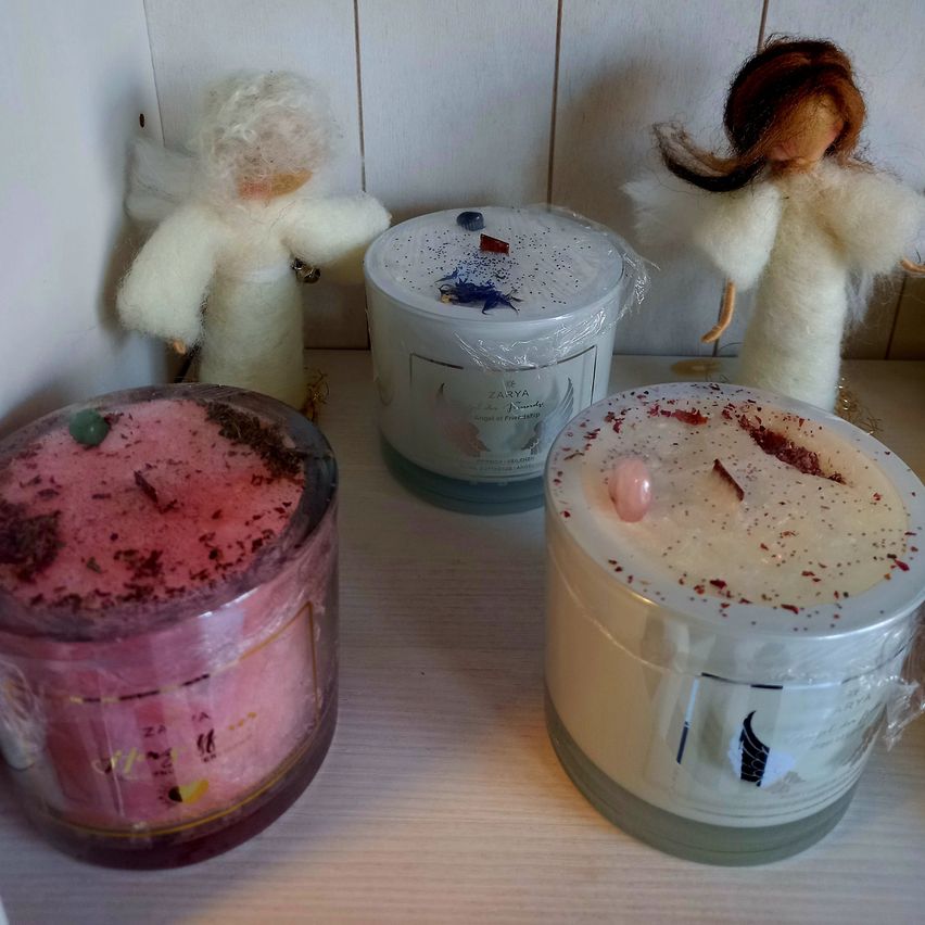 Drei Kerzen, zwei mit Deckeln, und zwei Engelsfiguren. Eine Kerze hat rosa Wachs und rote Blütenblätter oben, während die andere weißes Wachs und blaue Blütenblätter hat. Beide Kerzen haben Plastikdeckel.