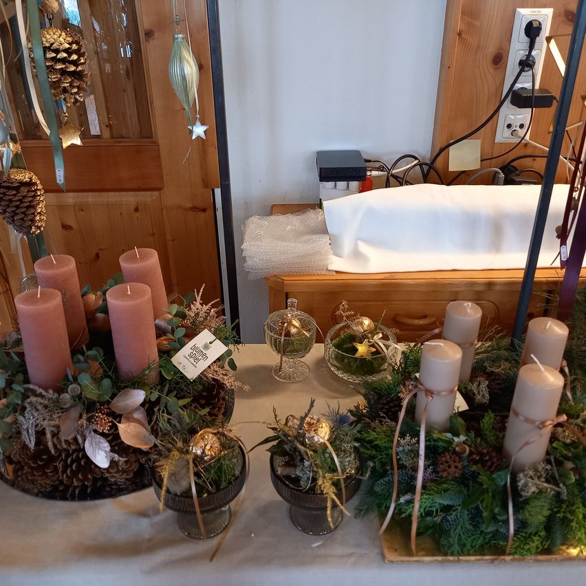 A table displays several candle arrangements, each with a different decoration, including pine cones, leaves, and ribbons. Behind the table, a wooden door and wall are visible, along with a power outlet and some items on a shelf.