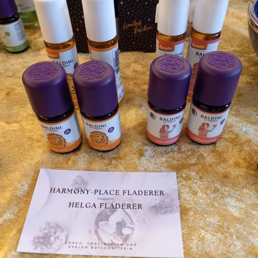 A collection of essential oils from Baldini on a gold surface, including bottles labeled 'Baldini' and 'Herzöffner'. A business card titled 'Harmony-Place Fladerer' is also present.