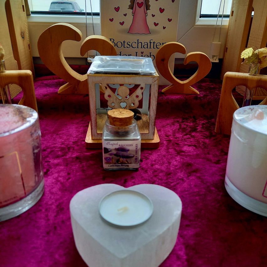 A heart-shaped candle sits on a table with other candles and wooden hearts. A small jar with a cork is beside it, and a poster with a pink angel is behind.