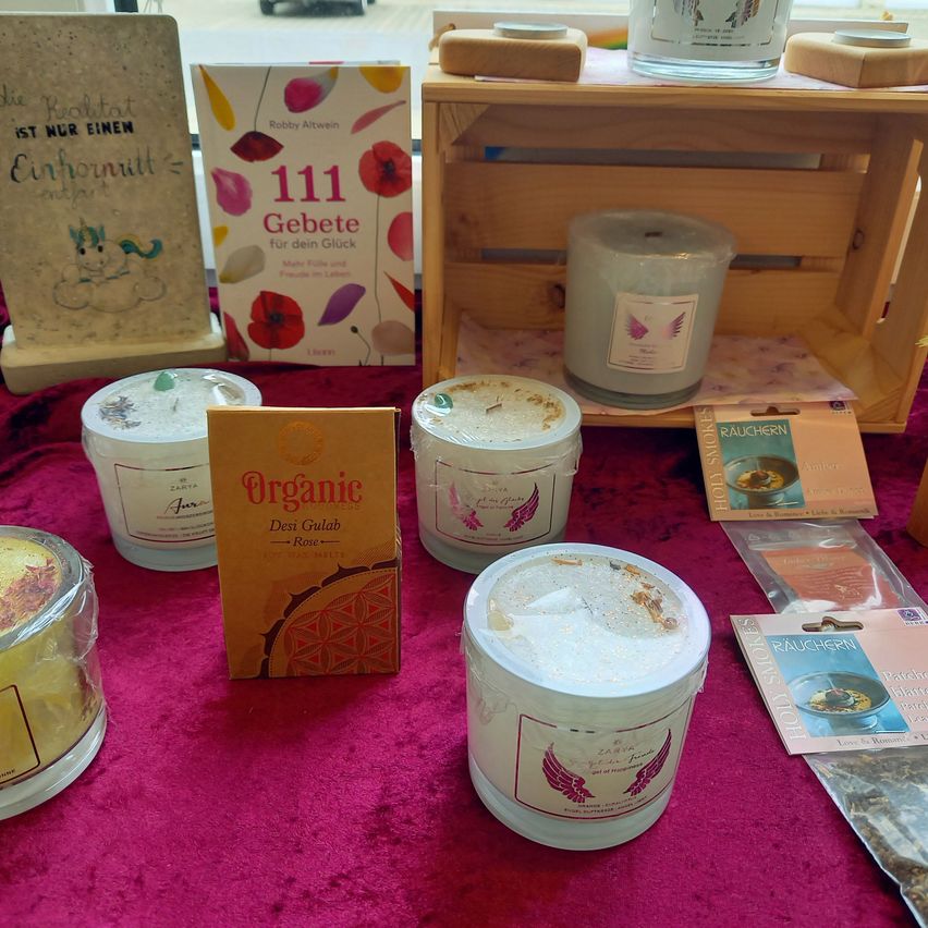 A wooden crate contains various items, including books, candles, and incense. Books on the table include '111 Gebete' and 'Die Realität ist nur ein Einhornritt'. Multiple candle jars with lids are on the table, along with other items.