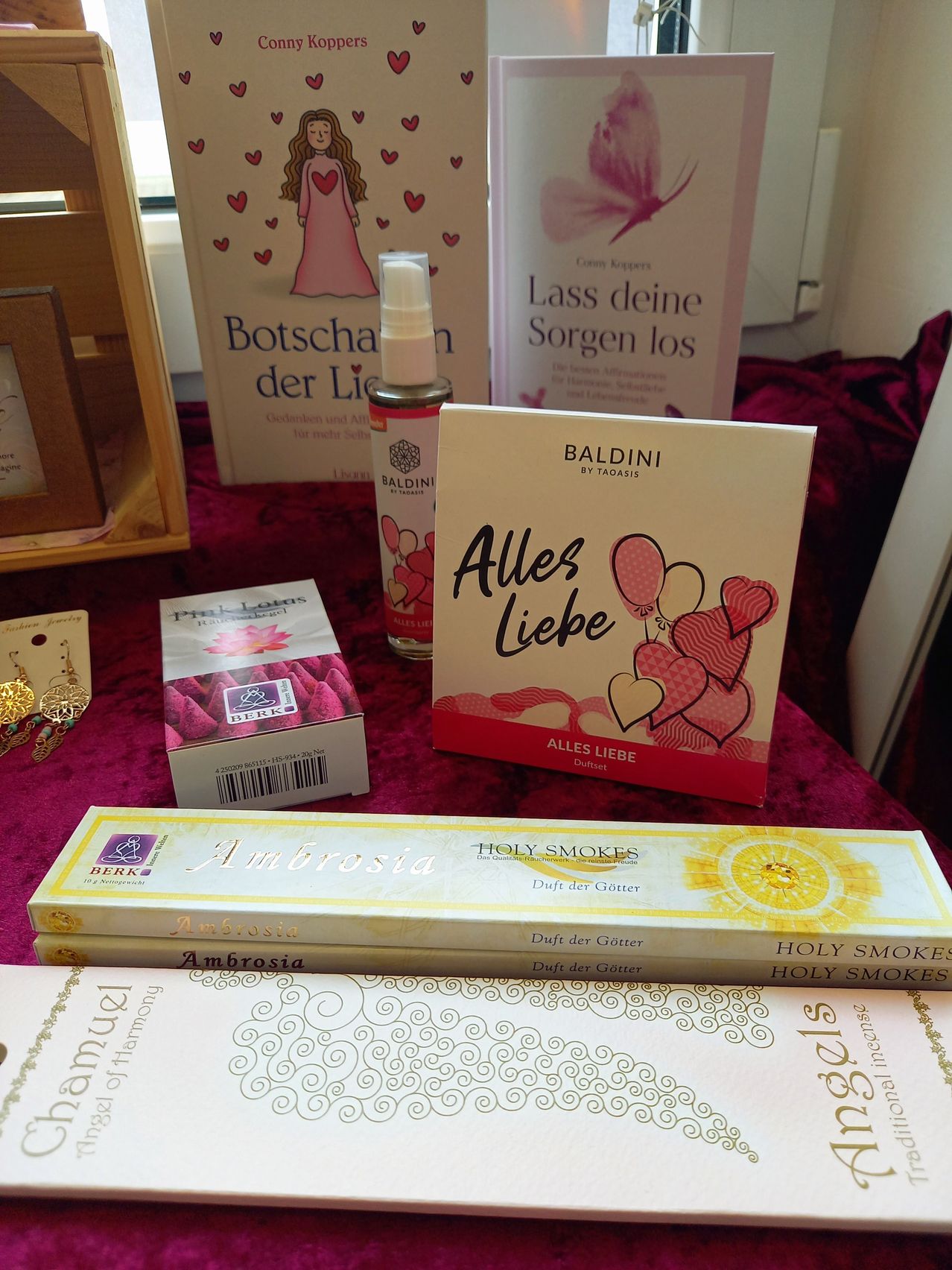 A table displays a variety of items: two boxes of incense, a box of sweets, a perfume bottle, and several books. The books have titles such as 'Botschaft der Liebe' and 'Lass deine Sorgen los'. The incense boxes are labeled 'Ambrosia' and 'Holy Smokes'.
