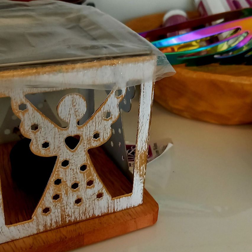 Eine hölzerne Engelsfigur mit Herz und Flügeln, teilweise von einer Plastikfolie bedeckt, auf einem Holzständer. Dahinter ein hölzerne Schüssel mit bunten Objekten.