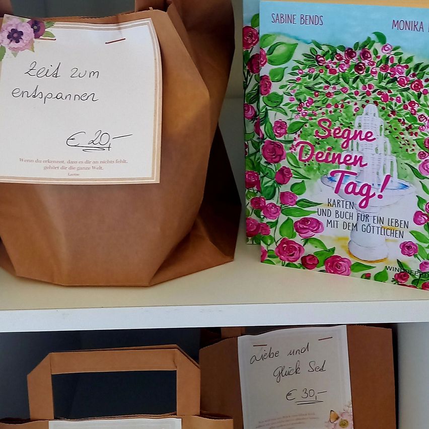 Ein brauner Papierbeutel mit einem weißen Etikett, auf dem 'Leis zum entspannen' steht, befindet sich auf einem Regal neben einem farbenfrohen Buch mit dem Titel 'Segne Deinen Tag!' und einem Blumendesign.