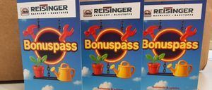 Drei Reisinger Bonuspass Werbekarten sind in einer Pappschachtel platziert. Die Karten zeigen farbenfrohe Werkzeuge und Pflanzen. Jede Karte hat einen blauen Hintergrund mit Text in gelber Schrift.