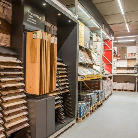Ein Innenbereich für Bodenbeläge zeigt verschiedene Holzbodenmuster, ordentlich auf Regalen gestapelt. Die Regale sind gut beleuchtet, und ein großes Schild mit der Aufschrift 'Bestellshop'. Neben den Regalen befinden sich weitere Produkte und eine Person in der Ferne.