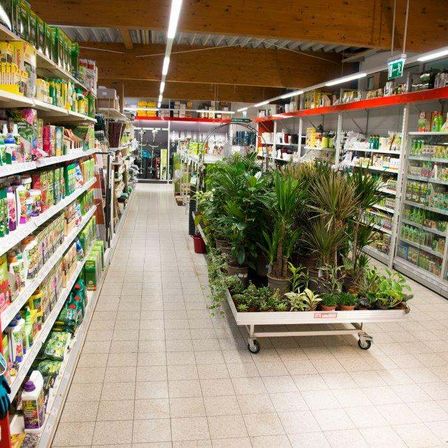 Ein Gang in einem Gartengeschäft mit weiß gefliestem Boden, Holzdecke und Regalen, die mit Gartenprodukten und Topfpflanzen gefüllt sind. In der Mitte befindet sich ein Trolley mit verschiedenen Pflanzen.