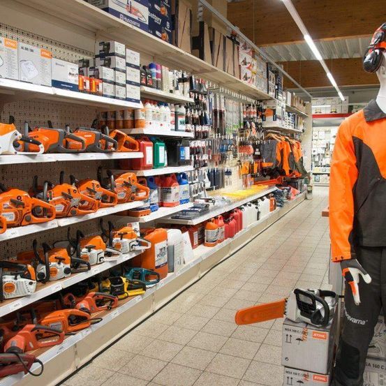 Ein Ladengang mit Reihen von Kettensägen in den Regalen. Schutzausrüstung wird an einem Mannequin ausgestellt. Verschiedene Produkte und Werkzeuge sind in den Regalen.