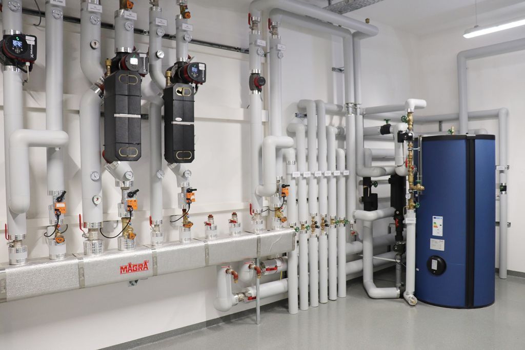 Ein Industrieraum mit weißen Rohren und Ventilen, die an einer weißen Wand befestigt sind, einem großen blauen Tank auf der rechten Seite und einem silbernen Rohr mit dem Wort 'MAGRA' darauf.