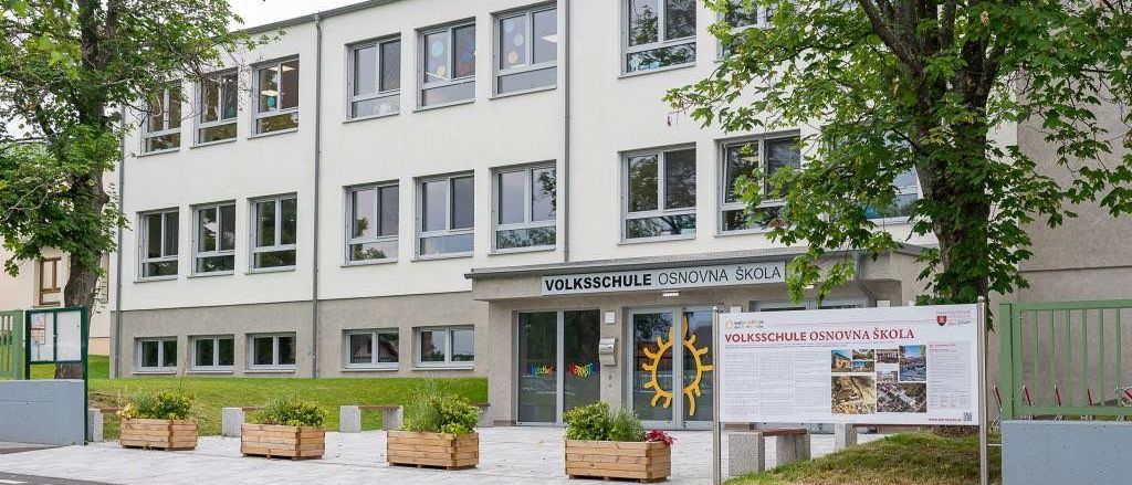 Die Vorderseite eines Erwachsenenbildungszentrums mit mehreren Fenstern und einem Schild, das 'Volksschule Osnovna Skole' lautet. Ein Plakat ist an einem Pfosten vor dem Eingang befestigt.