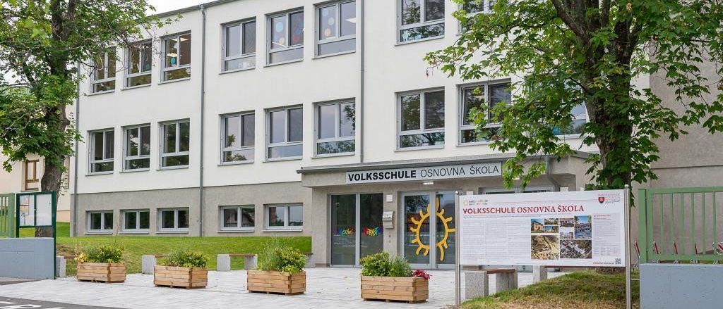 Ein modernes weißes Gebäude mit mehreren Fenstern, beschriftet mit 'VOLKSSCHULE OSNOVNA SKOLA', steht neben einem Baum. Davor befindet sich ein Schild mit Bildern. Mehrere Topfpflanzen stehen an der Seite.