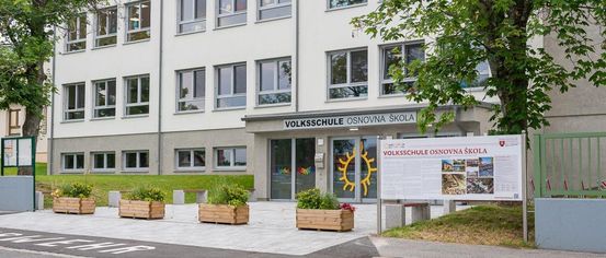 Die Vorderseite eines weißen Gebäudes mit Glasfenstern und einem Schild, das Volksschule Osnovna Skole besagt. Der Eingang hat eine Tür mit einem Sonnensymbol.