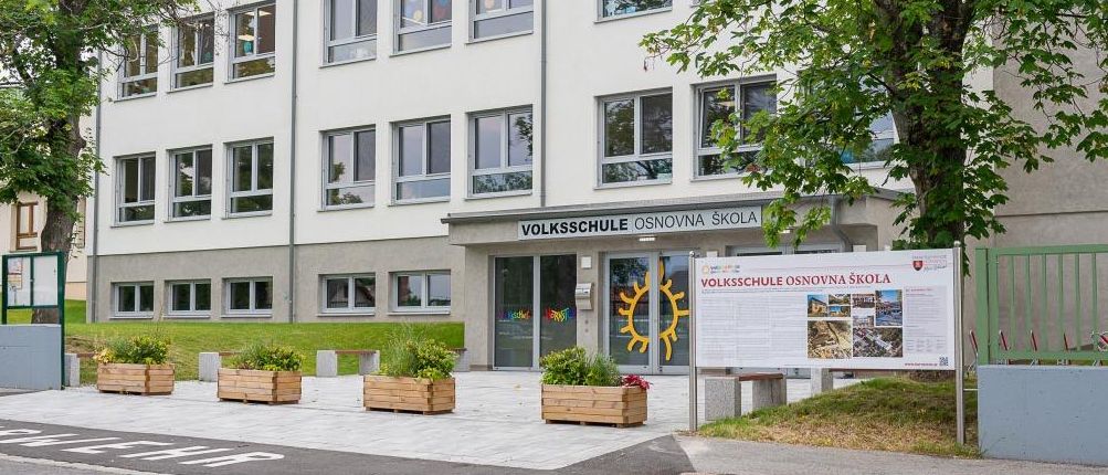 Die Vorderseite eines weißen Gebäudes mit Glasfenstern und einem Schild, das Volksschule Osnovna Skole besagt. Der Eingang hat eine Tür mit einem Sonnensymbol.