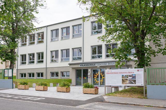 Der Eingang der Volksschule Osnovna Skole, ein Schulgebäude mit mehreren Glasfenstern, einem gelben Sonnenlogo und einem Schild. Davor befinden sich Bäume, Topfpflanzen und ein grüner Zaun.