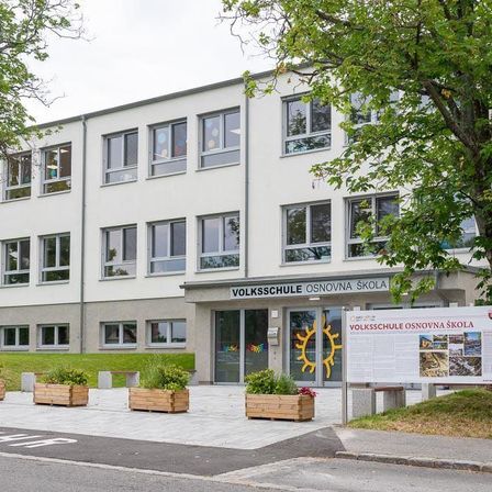 Ein weißes Gebäude mit mehreren Fenstern, beschriftet als 'Volksschule Osnovna skola', mit einem großen Schild und einem gelben Sonnenlogo.