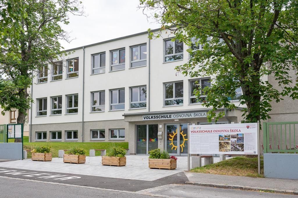 Ein weißes Gebäude mit mehreren Fenstern, beschriftet als 'Volksschule Osnovna skola', mit einem großen Schild und einem gelben Sonnenlogo.