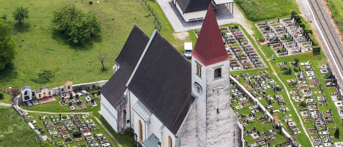 Ein Luftbild einer Kirche mit einem roten Turm, umgeben von einem Friedhof mit vielen Grabsteinen und Pflanzen. Ein Weg führt zur Kirche.