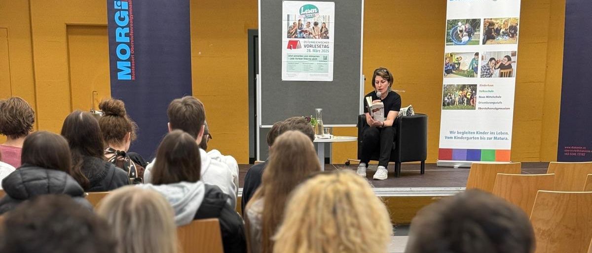 Studenten sitzen in Reihen in einem Hörsaal, eine Frau sitzt auf der Bühne und liest aus einem Buch. Der Raum ist mit Menschen gefüllt und die Bühne hat einen Tisch mit einer Flasche und einem Stuhl.