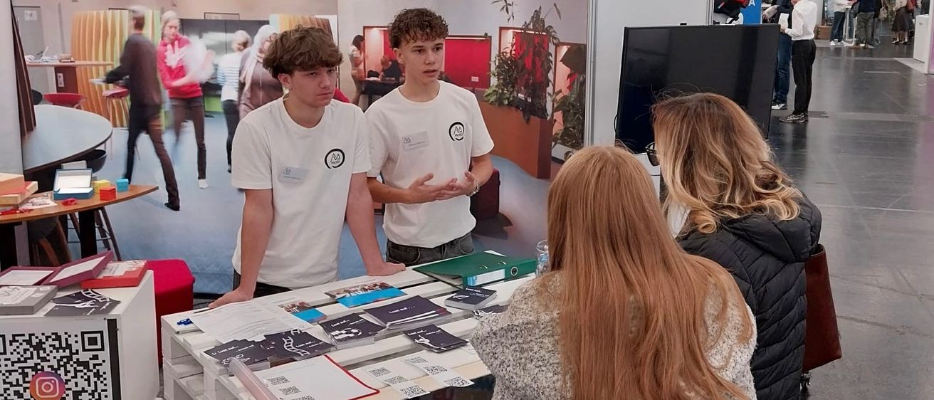 In einem Saal stehen zwei junge Männer hinter einem Tisch mit Broschüren und unterhalten sich mit zwei Frauen. Dahinter zeigt ein Banner ein Klassenzimmer. Im Hintergrund gehen Leute, und ein Tisch hält Bücher und einen Monitor.
