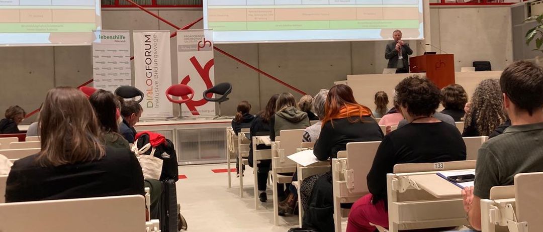 Ein Mann spricht an einem Podium mit einer Projektionsleinwand hinter ihm. Ein Publikum sitzt in Stühlen vor ihm.